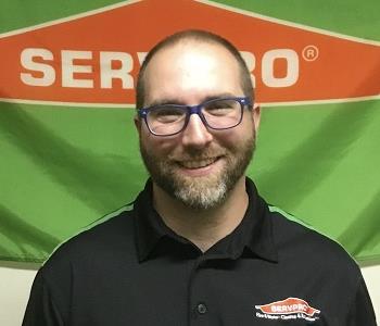 bearded male employee with glasses standing in front of a SERVPRO logo'd banner